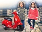 Dwoje dzieci pozuje obok czerwonego dziecięcego skutera Vespa.