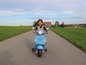 Dziewczynka jedzie na niebieskim skuterze Vespa po szutrowej drodze.