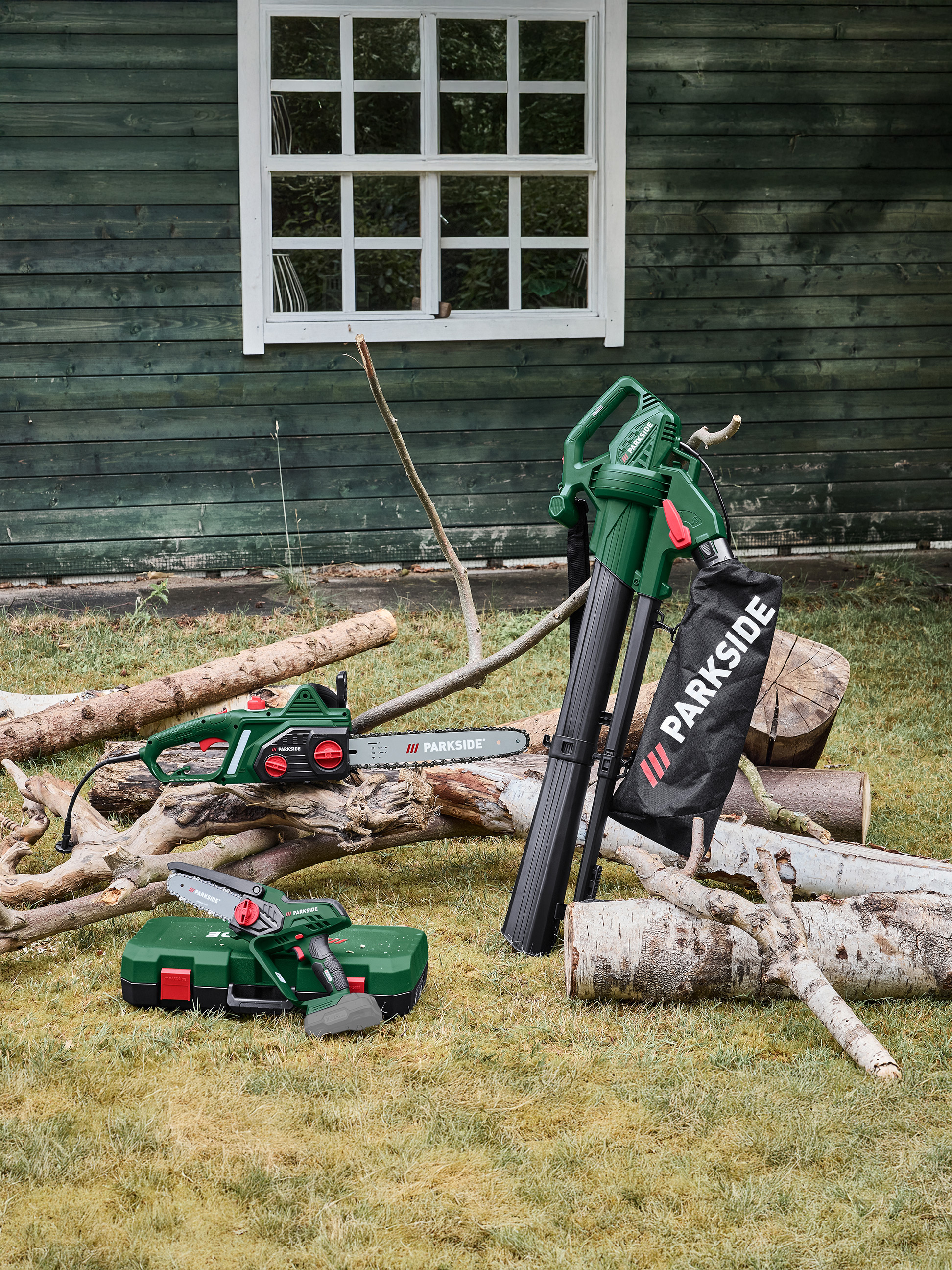 Dmuchawa do liści, piła łańcuchowa i mini piła łańcuchowa na trawie obok drewnianej szopy.