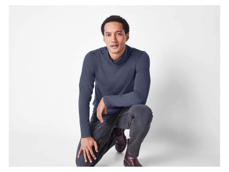 Mężczyzna w szarym golfie i szarych dżinsach, kucający na białym tle.