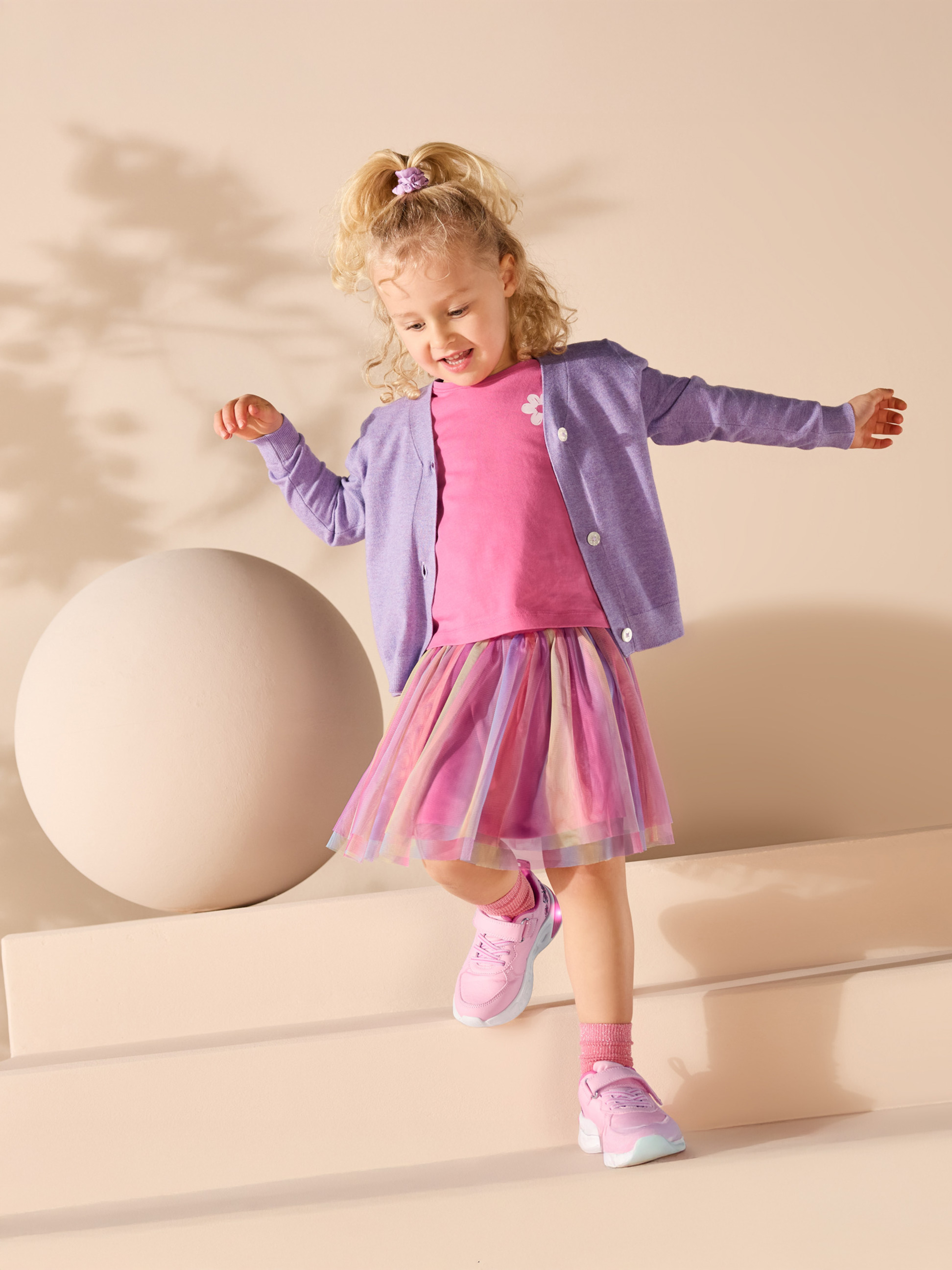 Dziewczynka w różowej koszulce, fioletowym kardiganie i kolorowej tiulowej spódnicy.