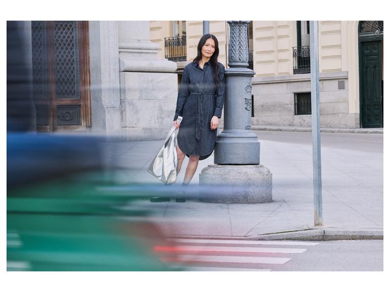 Kobieta w dżinsowej sukience stoi przy słupku, trzymając srebrną torbę.