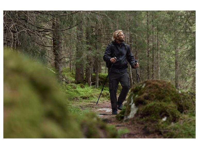 Mężczyzna z długimi blond włosami w czarnej kurtce i spodniach cargo, wędrujący z kijkami.
