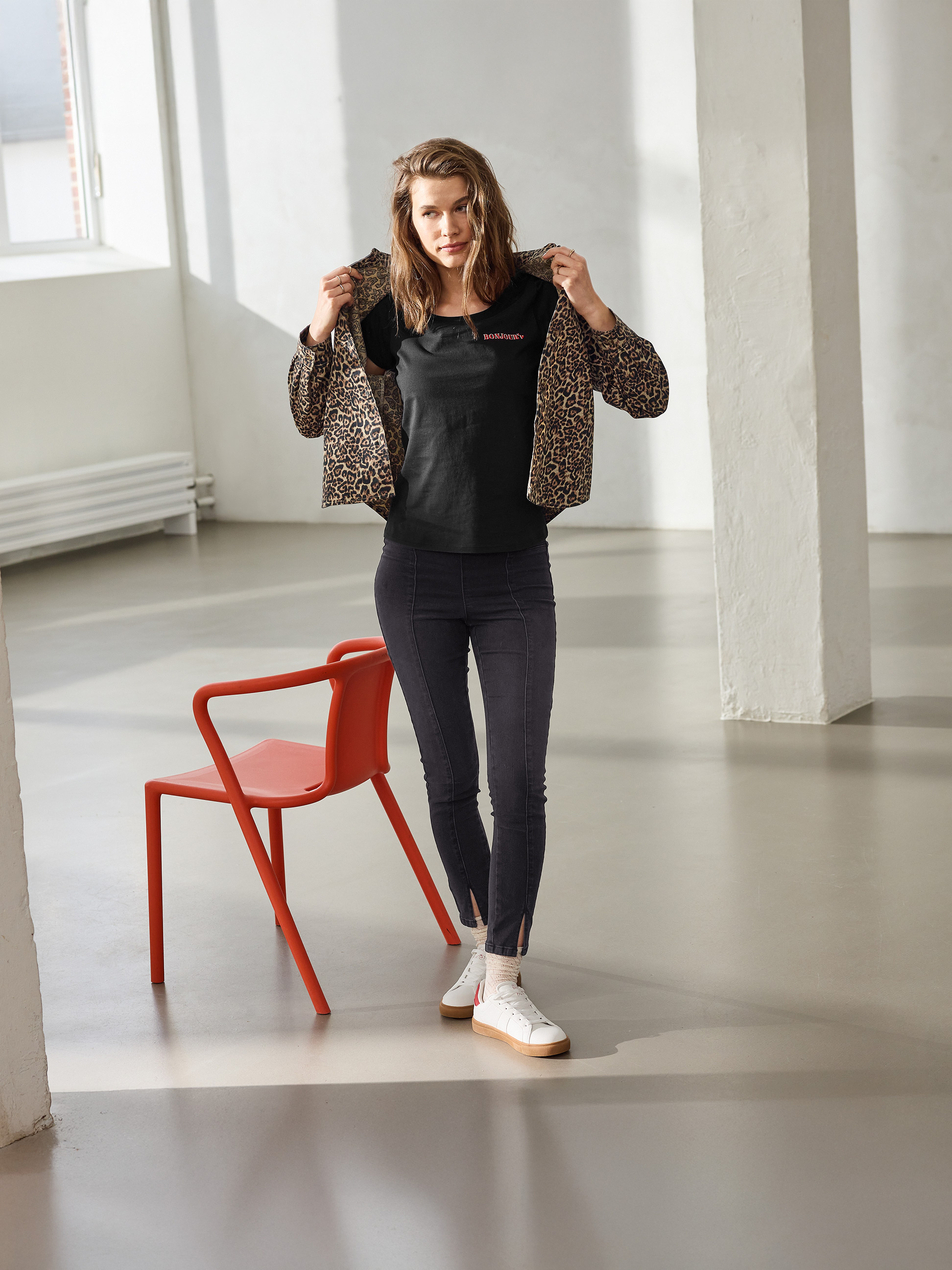 Kobieta w kurtce w panterkę, czarnym T-shircie 'BONJOUR' i czarnych obcisłych dżinsach, pozująca obok czerwonego krzesła.