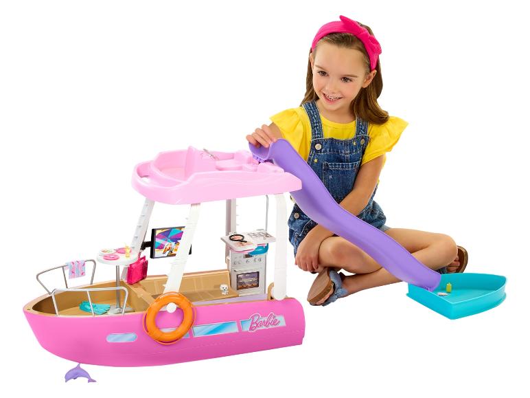Uśmiechnięta dziewczynka bawi się różową łodzią-zabawką Barbie, ze zjeżdżalnią i akcesoriami.