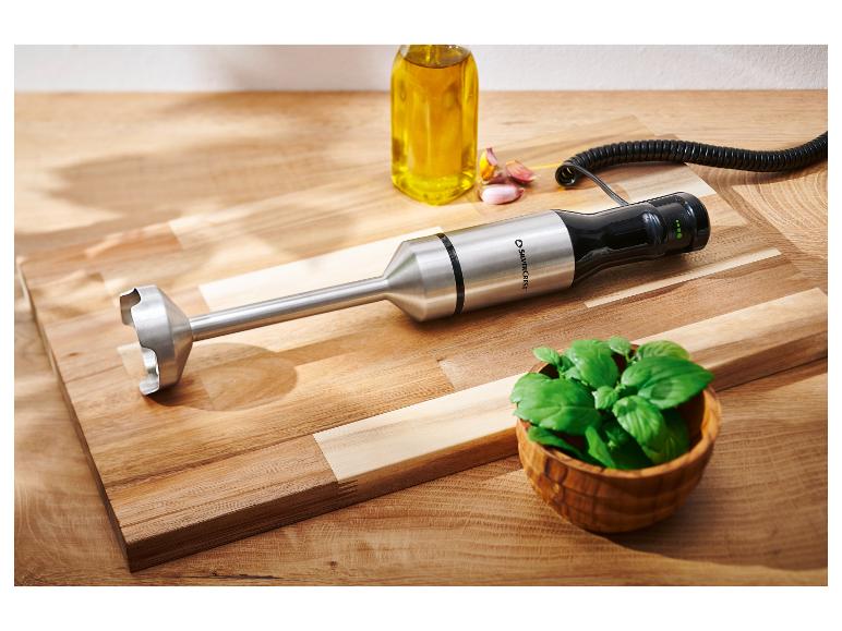 Blender ręczny, butelka oleju, czosnek i bazylia na drewnianej desce.