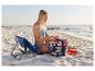Kobieta w bikini na plaży, siedząca obok niebieskiego krzesła plażowego i torby z tropikalnym wzorem.