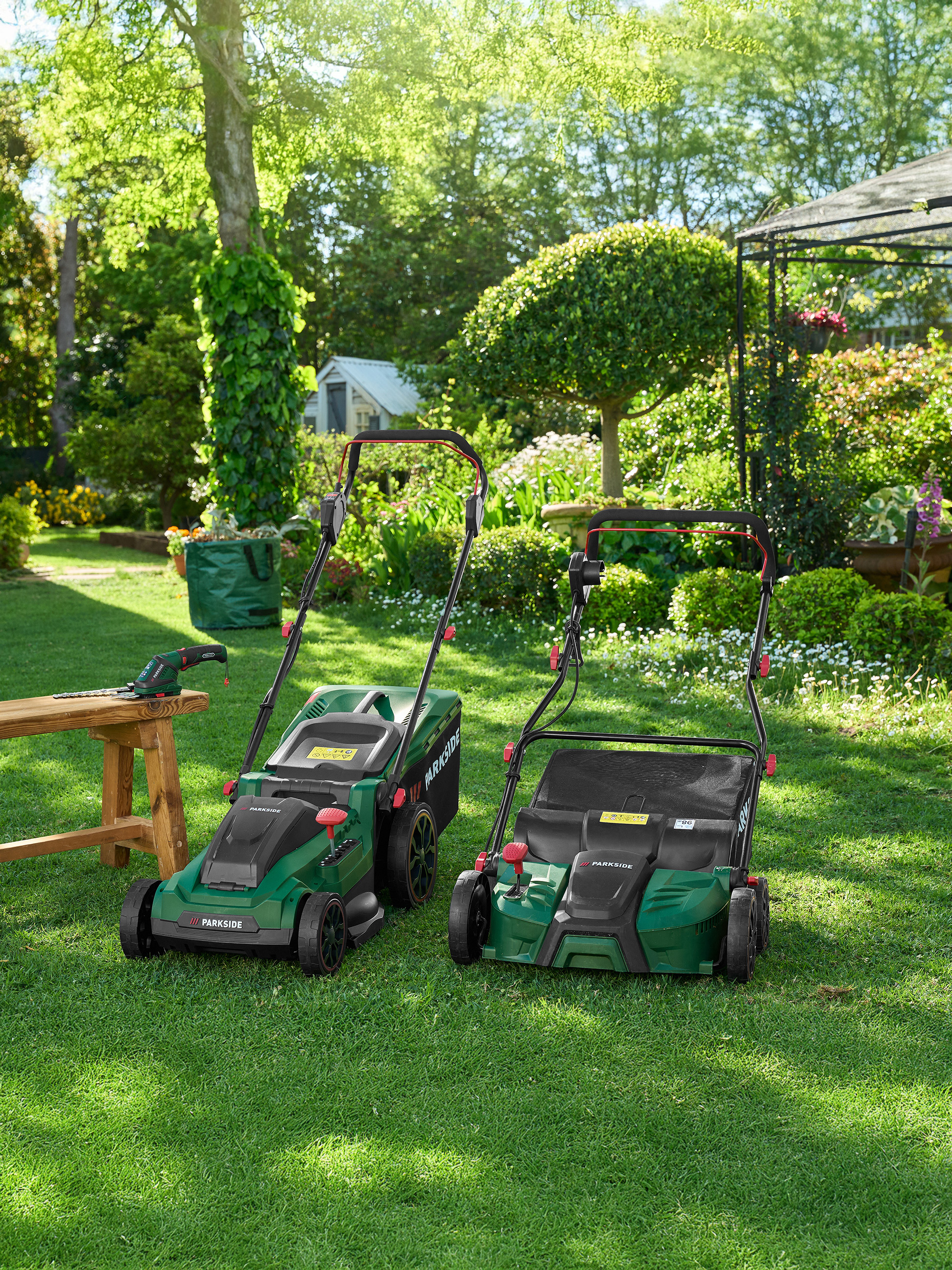 Dwie zielone kosiarki Parkside i narzędzia ogrodowe na trawniku w ogrodzie.