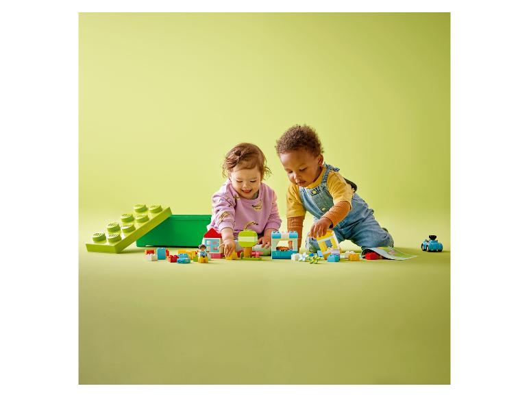 Dwoje małych dzieci bawi się kolorowymi klockami i figurkami LEGO DUPLO na zielonej powierzchni.