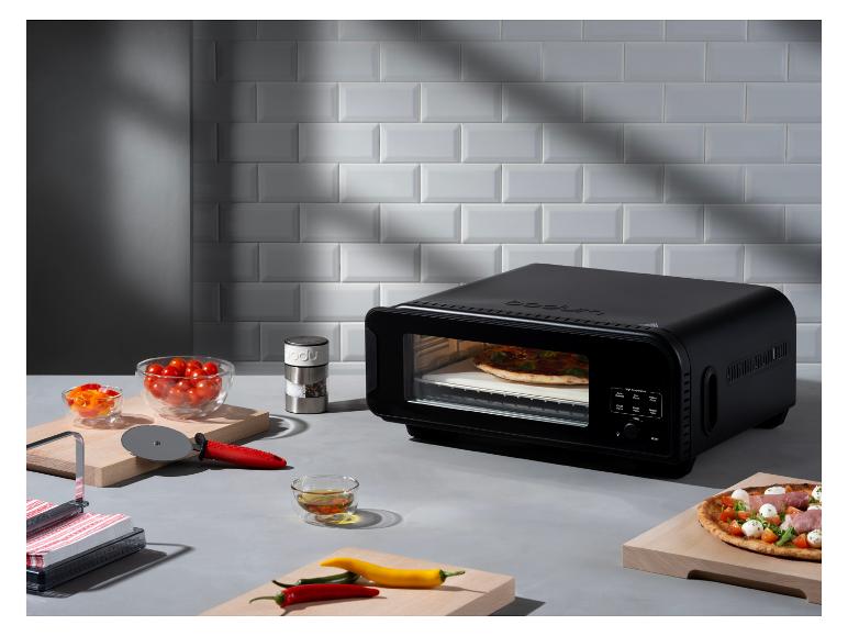 Czarny piec do pizzy z pizzą w środku, otoczony świeżymi składnikami i narzędziami kuchennymi.