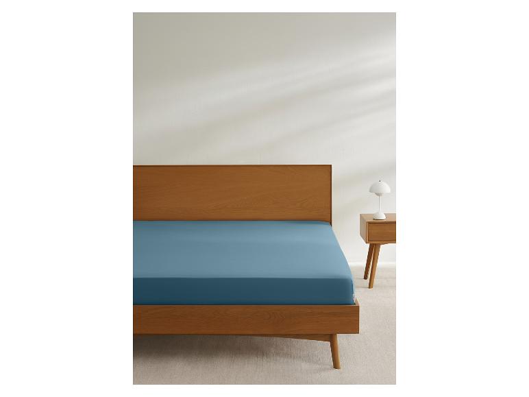 Łóżko z drewnianą ramą i niebieskim prześcieradłem, obok szafka nocna z lampką.