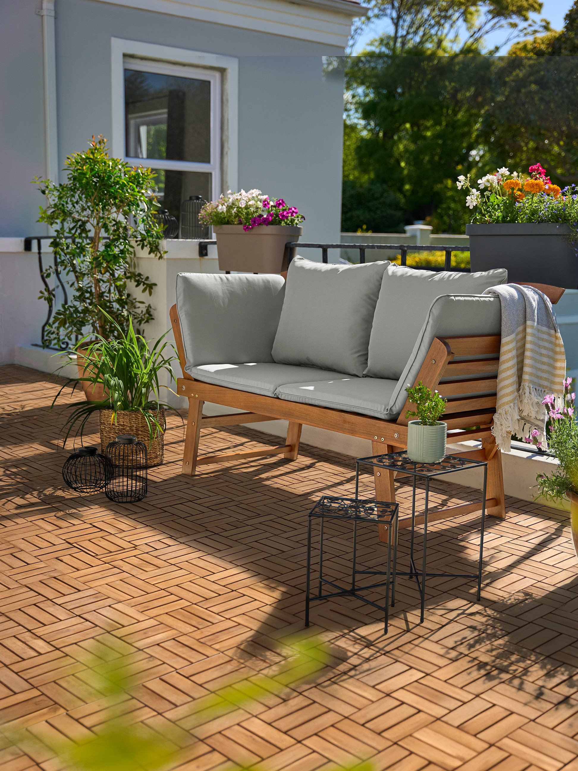 Drewniana sofa ogrodowa z szarymi poduszkami na balkonie z drewnianymi płytkami podłogowymi i roślinami