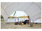 Kobieta siedzi pod plandeką z kocem, podczas gdy inne osoby bawią się na plaży w oddali.