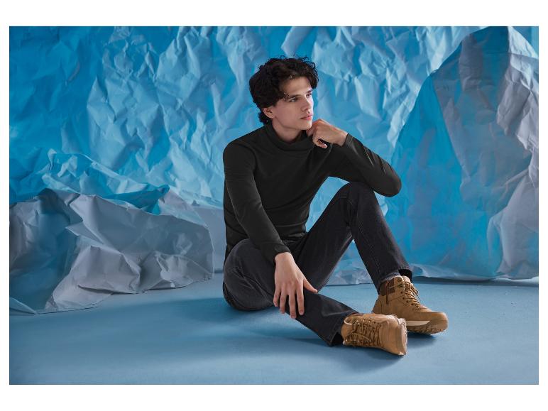 Mężczyzna w czarnym golfie i dżinsach siedzi na podłodze w brązowych butach.