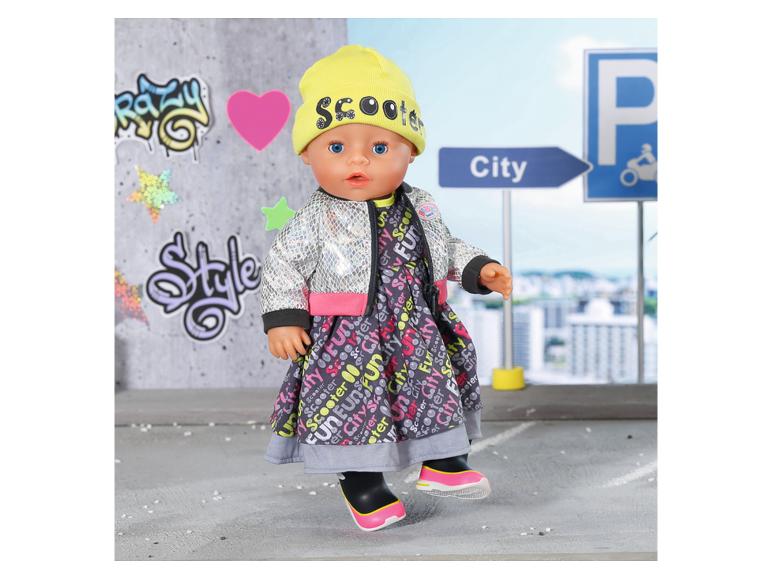 Lalka w sukience z miejskim wzorem, srebrnej kurtce i żółtej czapce z napisem „Scooter”.