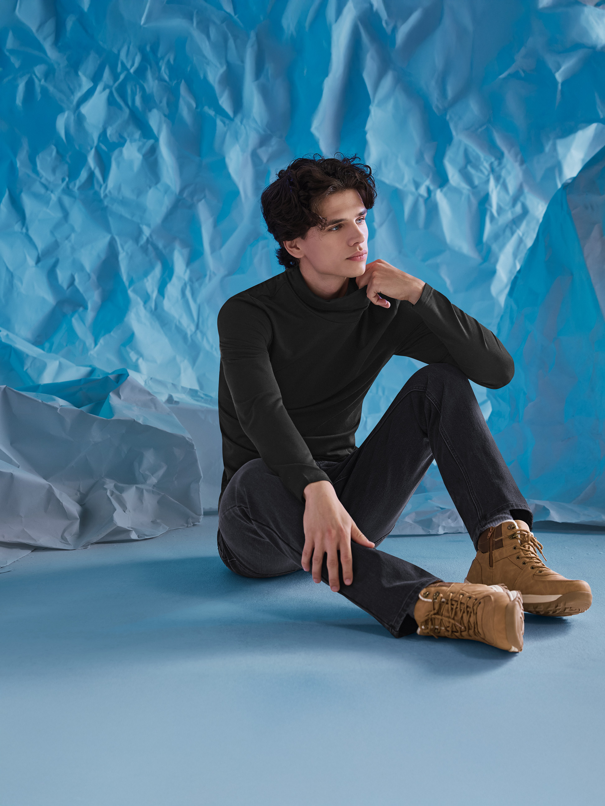 Mężczyzna w czarnym golfie, dżinsach i brązowych butach, siedzący na niebieskim tle.