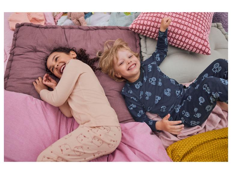 Dzieci śmieją się, leżąc w łóżku z pościelą w pastelowych kolorach i wzorach z kotami i traktorami.