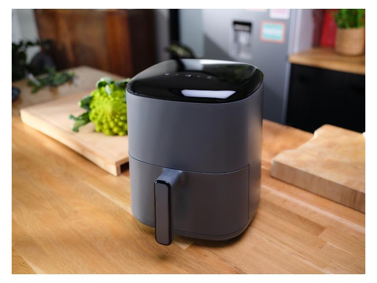 Szara frytkownica beztłuszczowa na drewnianym blacie kuchennym, z warzywami w tle.