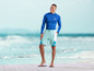 Mężczyzna w niebieskiej koszulce rashguard i niebieskich spodenkach kąpielowych z logo 'mi' na plaży.