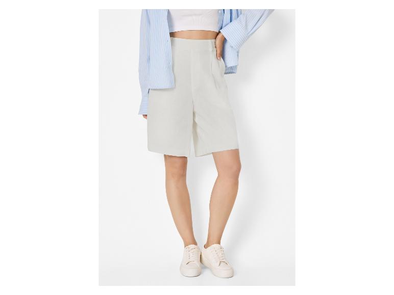 Kobieta w jasnoszarych szortach, białym crop topie, niebieskiej koszuli w paski i białych sneakersach.