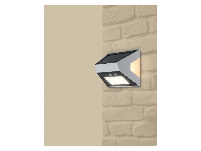 Lampa solarna ścienna z czujnikiem ruchu i dwoma światłami, zamontowana na ceglanej ścianie.