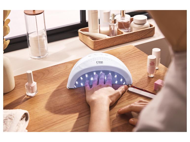Kobieta suszy paznokcie w lampie UV, obok lakiery i akcesoria do manicure.