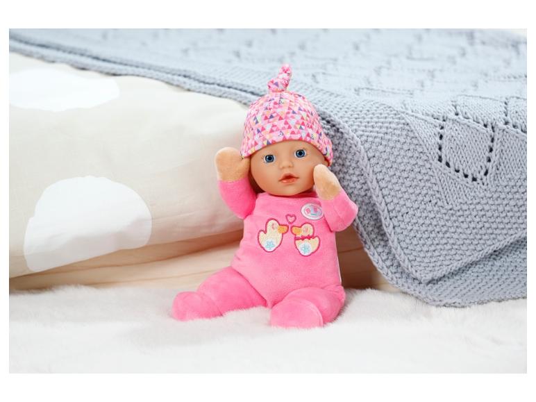 Lalka Baby Born w różowym ubranku i czapce na białym pluszowym dywanie.