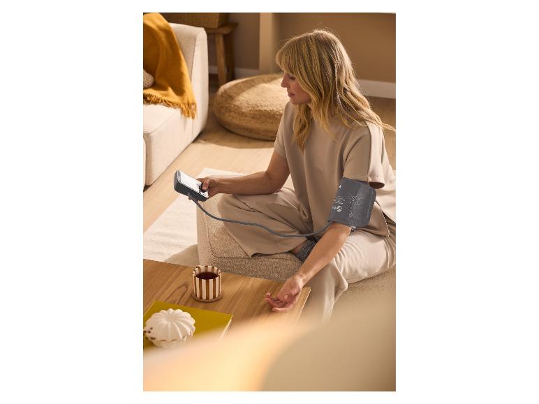 Kobieta mierząca ciśnienie krwi za pomocą naramiennego ciśnieniomierza, siedząca na podłodze z filiżanką herbaty.