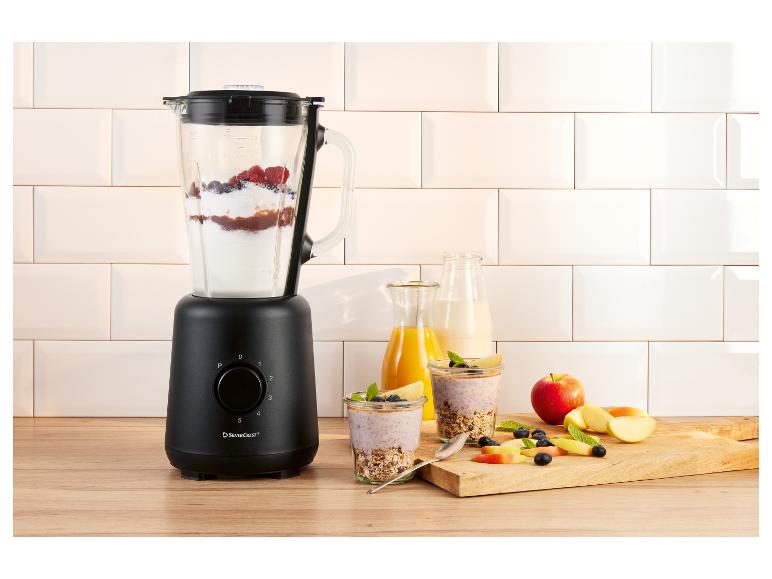 Czarny blender z jagodami i jogurtem, obok szklanek ze śniadaniem i owocami.