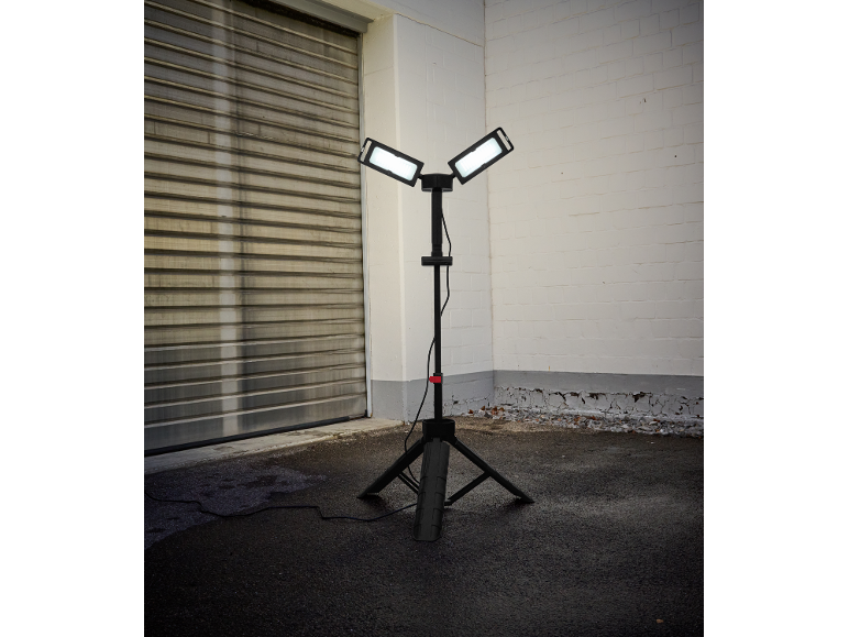 Lampa robocza na statywie z dwoma podświetlanymi panelami na asfalcie, obok bramy garażowej i ceglanej ściany.