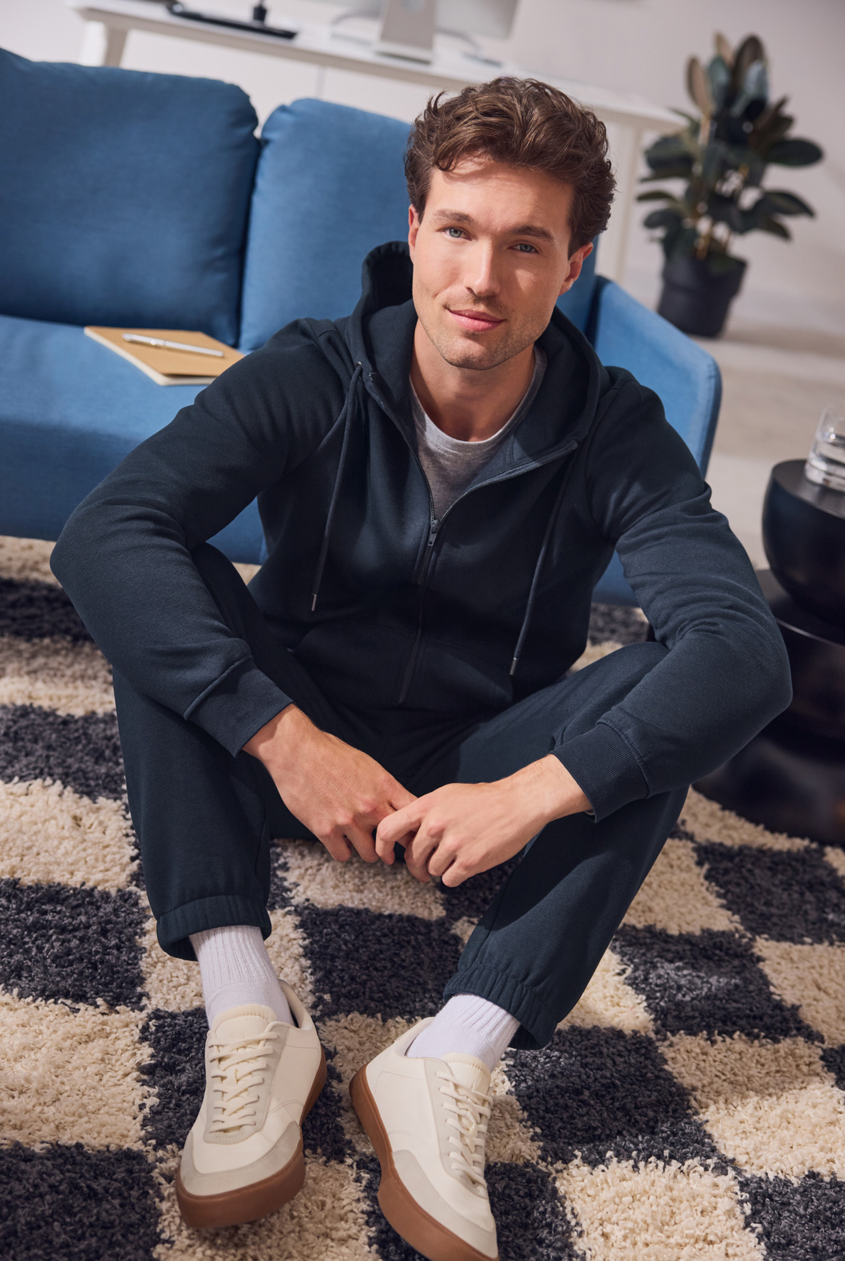 Mężczyzna w granatowej bluzie z kapturem i spodniach dresowych siedzi na dywanie w kratę.