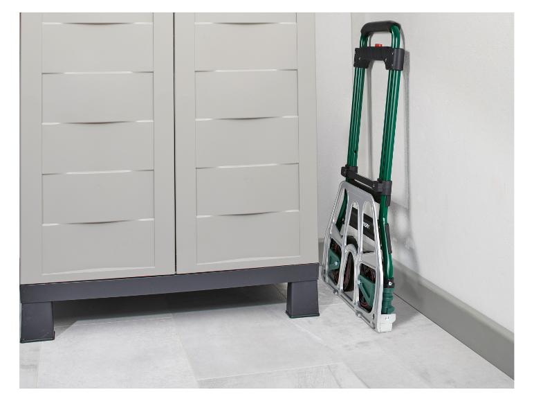 Złożony zielono-srebrny wózek ręczny oparty o białą ścianę obok jasnoszarej szafki do przechowywania.