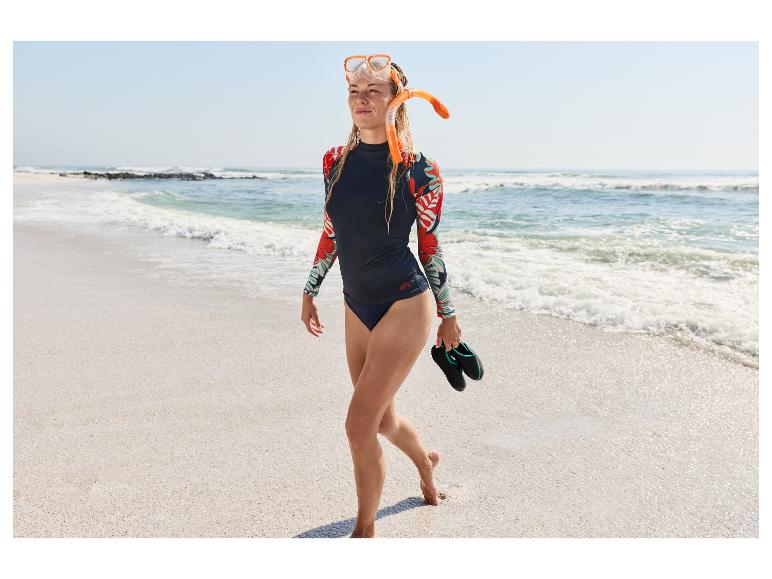 Kobieta w zestawie do snorkelingu CRIVIT i koszulce UV idzie po piaszczystej plaży.
