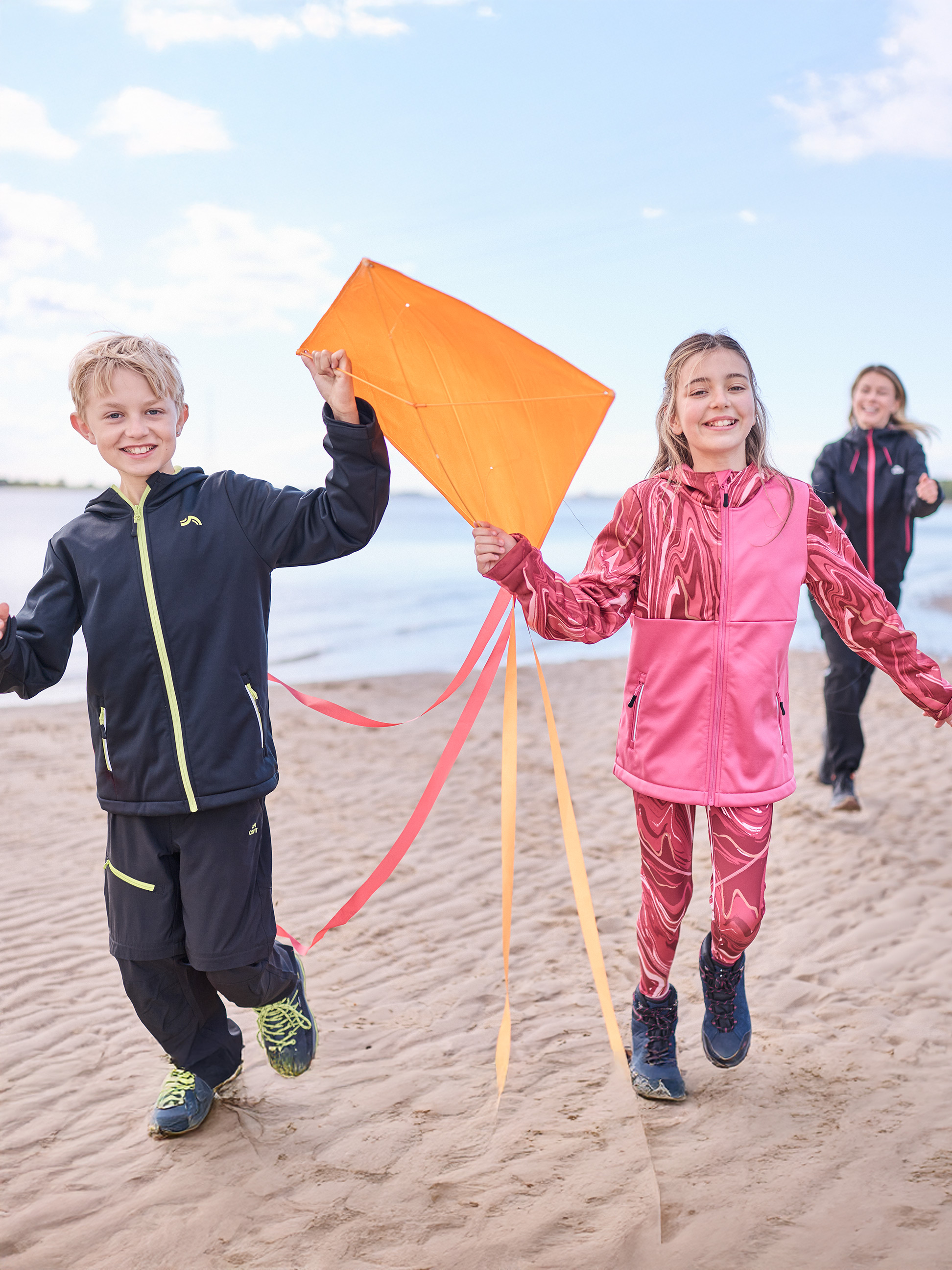 Dzieci w odzieży sportowej biegną po plaży z latawcem, kobieta w tle.