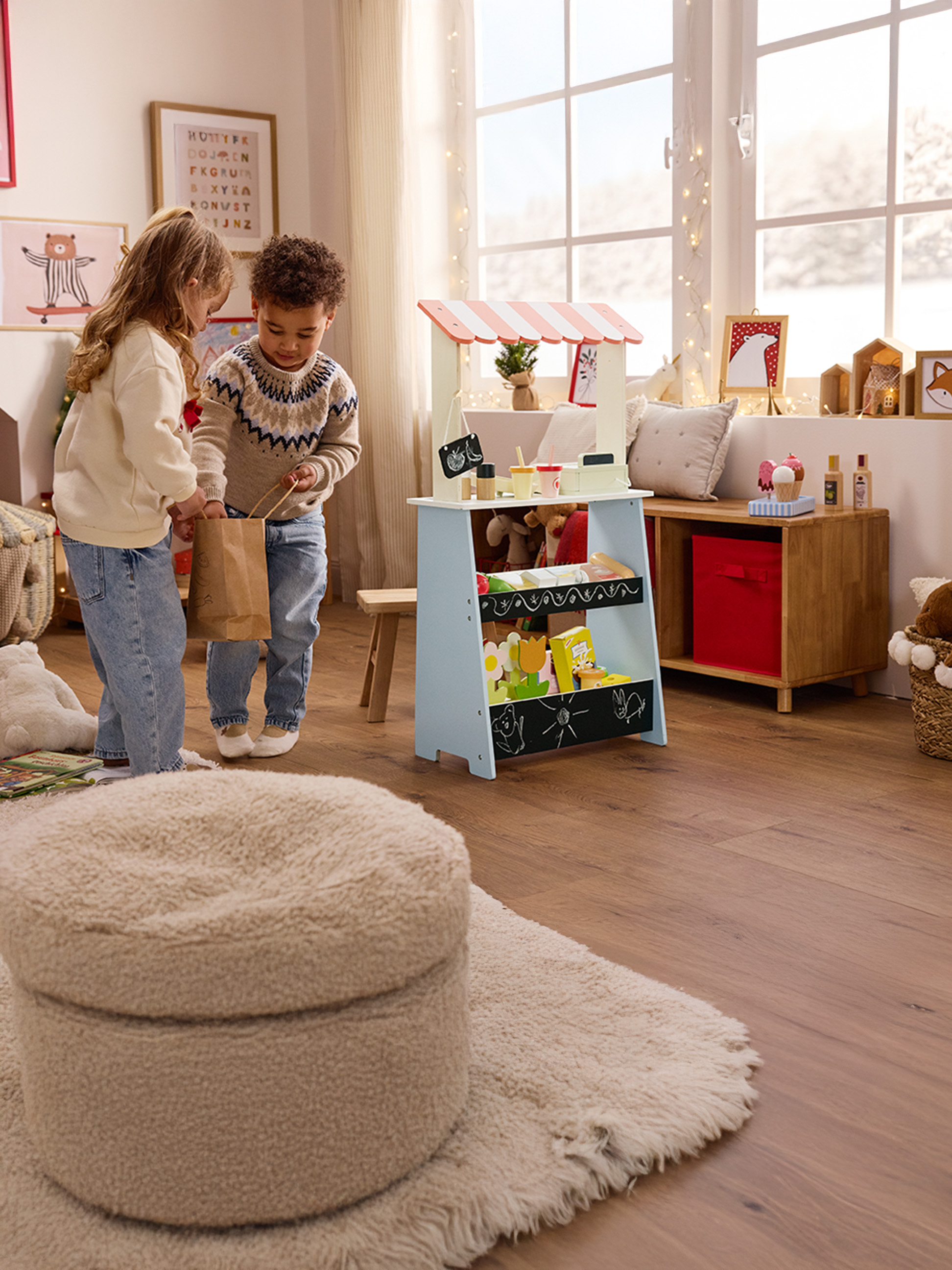 Dwoje dzieci bawi się drewnianym straganem, dziewczynka trzyma papierową torbę.