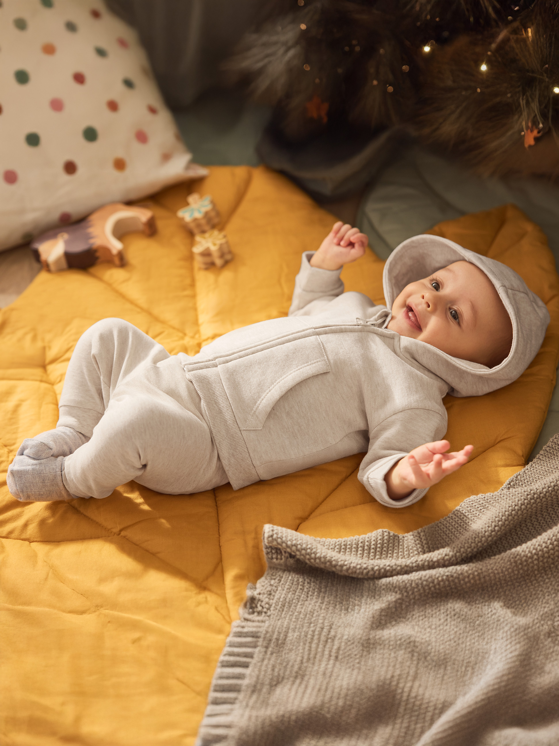 Szczęśliwe dziecko w szarym kombinezonie na żółtym materacu z zabawkami i lampkami choinkowymi.