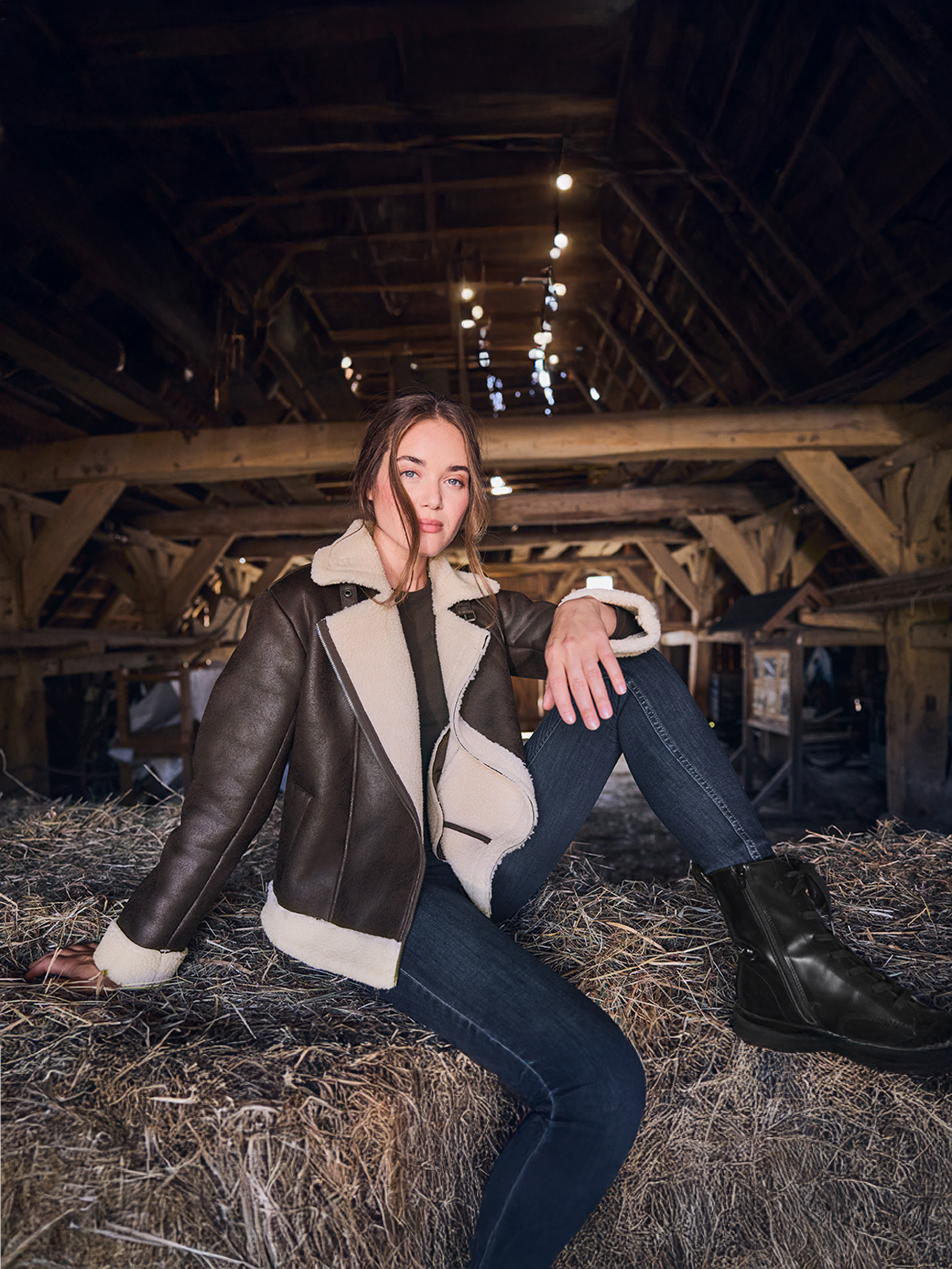 Kobieta w brązowej kurtce shearling i dżinsach, siedząca na belach siana w stodole.