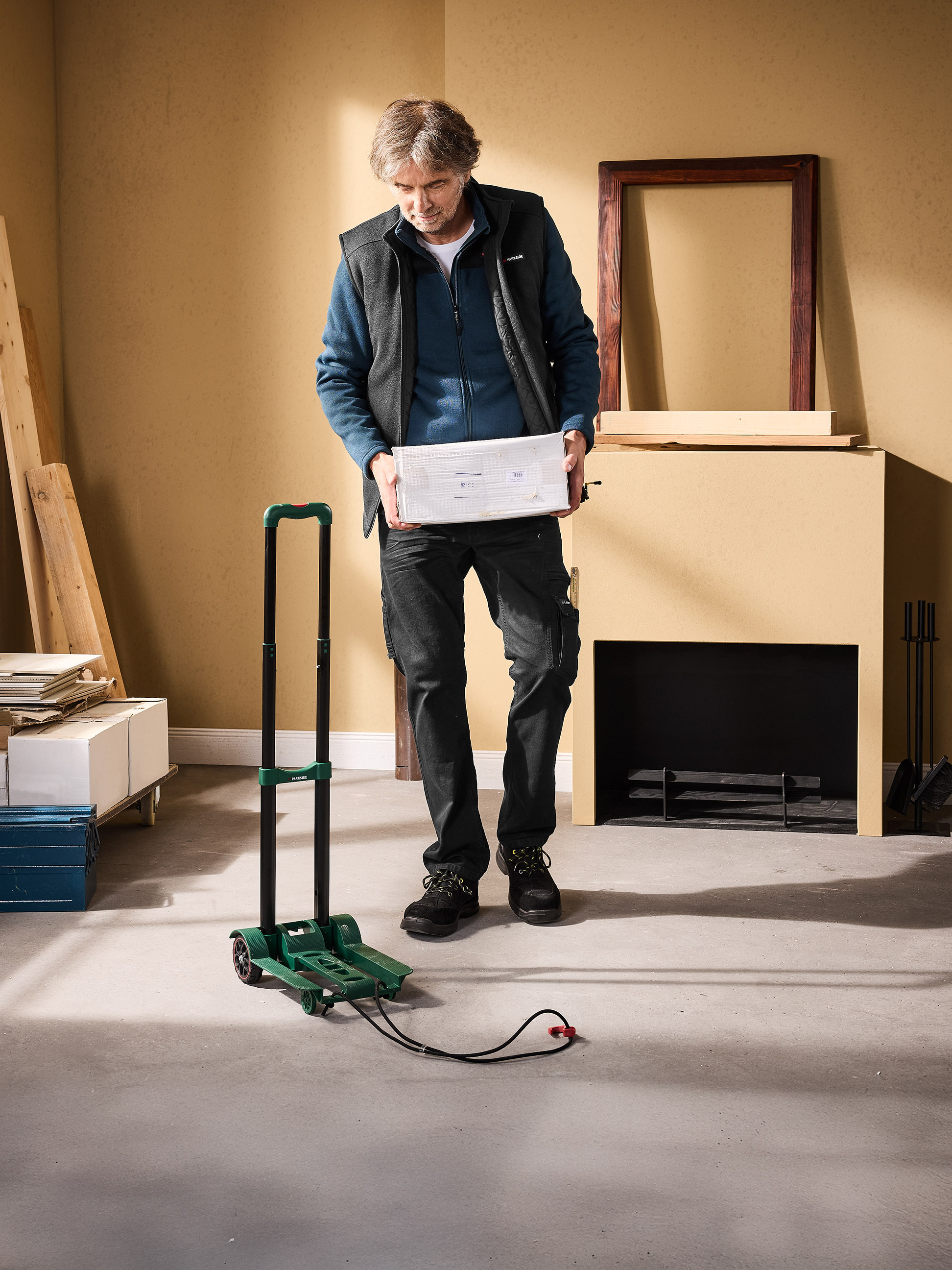 Mężczyzna trzyma białe pudełko, obok zielonego wózka i kominka.