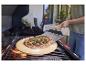 Pizza na kamieniu do pizzy na grillu, z osobą używającą łopaty do pizzy.