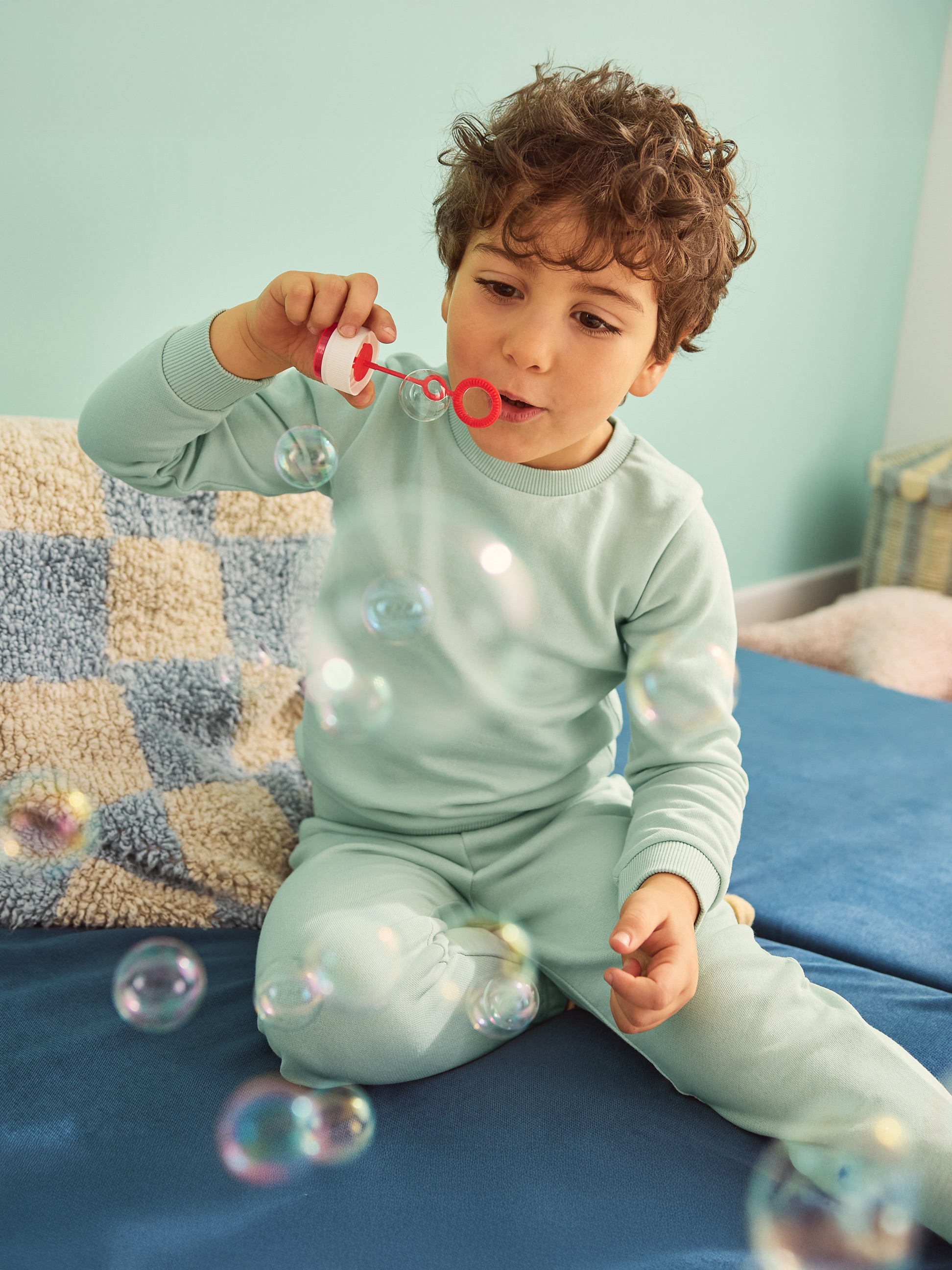 Chłopiec w niebieskiej piżamie dmucha bańki na niebieskiej sofie.