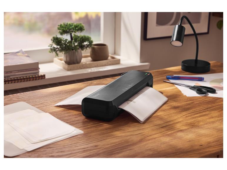 Czarny laminator na drewnianym biurku z papierami, nożyczkami i lampką biurkową.
