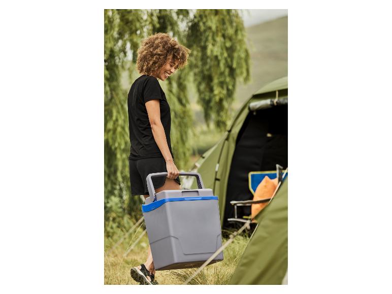 Uśmiechnięta kobieta niosąca szarą lodówkę turystyczną obok namiotu kempingowego.