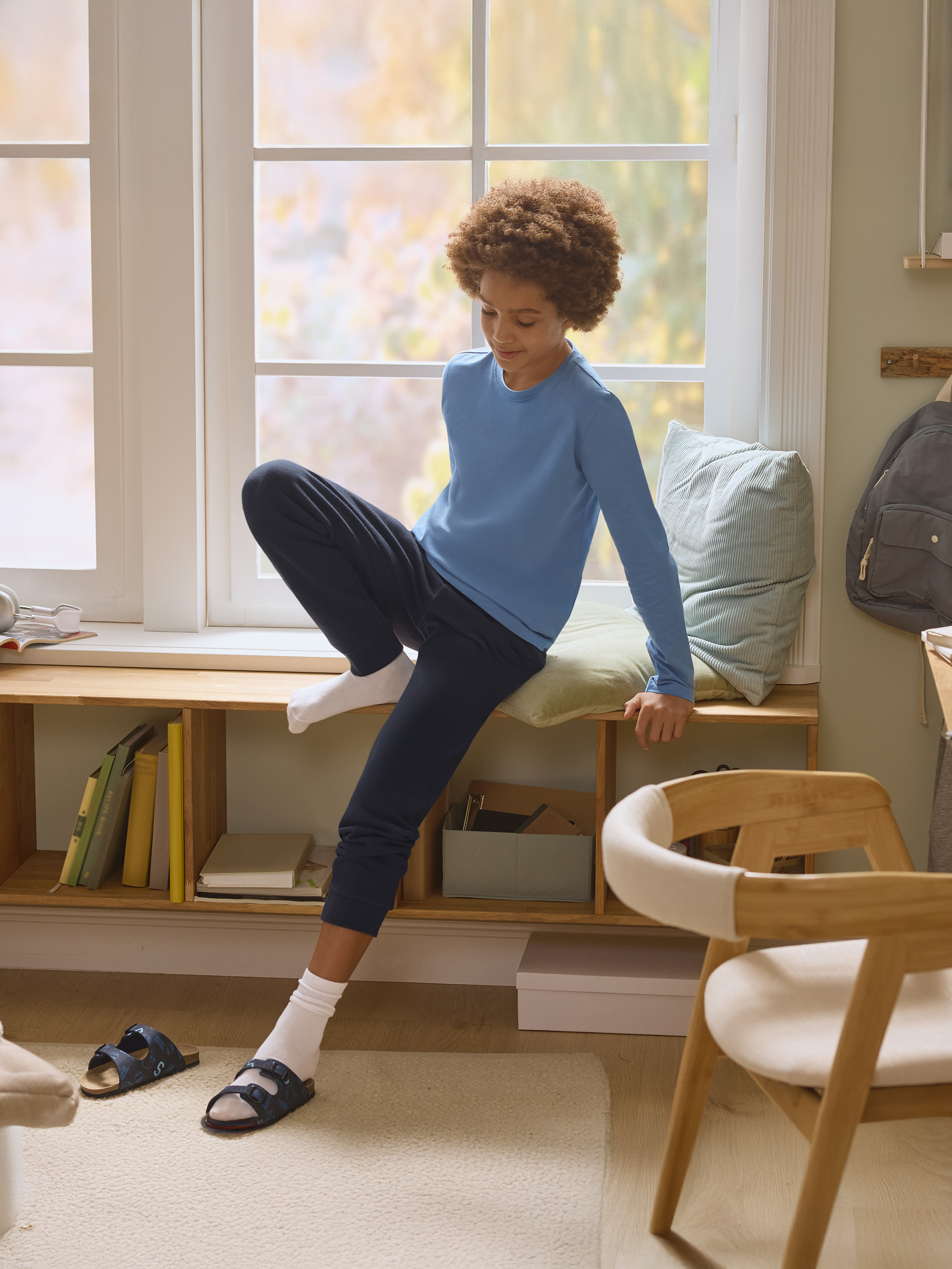 Chłopiec w niebieskiej piżamie siedzi na parapecie z książkami i poduszkami.