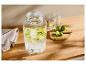 Szklany dozownik napojów z pokrojonymi limonkami, szklankami i całymi limonkami na drewnianym stole.