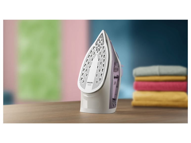 Białe żelazko Philips z ceramiczną płytą, na drewnianym stole ze stosem ubrań.