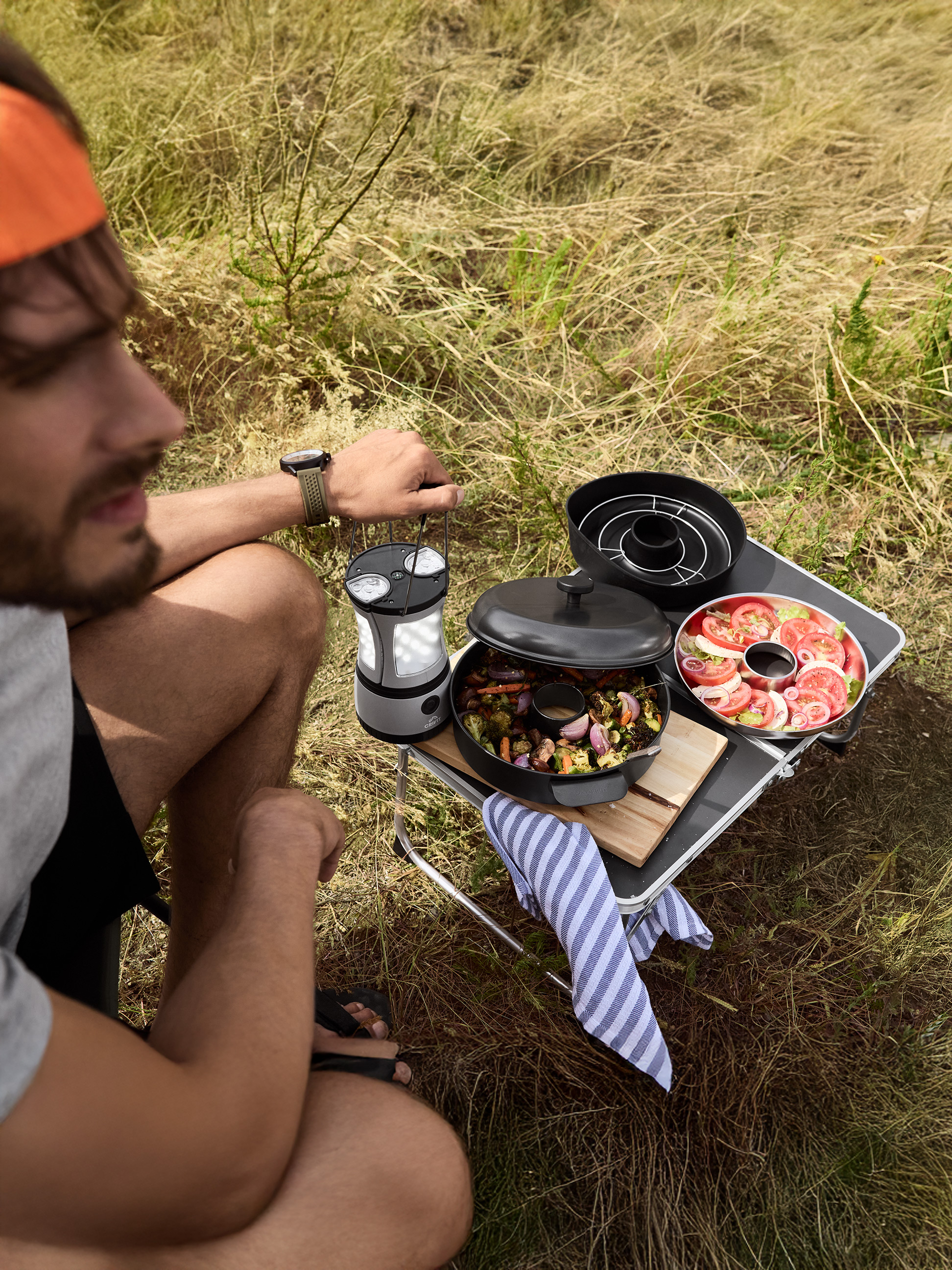 Mężczyzna gotuje na świeżym powietrzu z przenośnym grillem, serwując pieczone warzywa i sałatkę pomidorową.