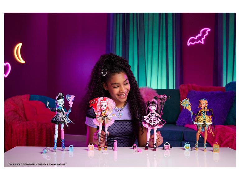 Uśmiechnięta dziewczyna trzyma lalki Monster High, z innymi lalkami i akcesoriami na stole.