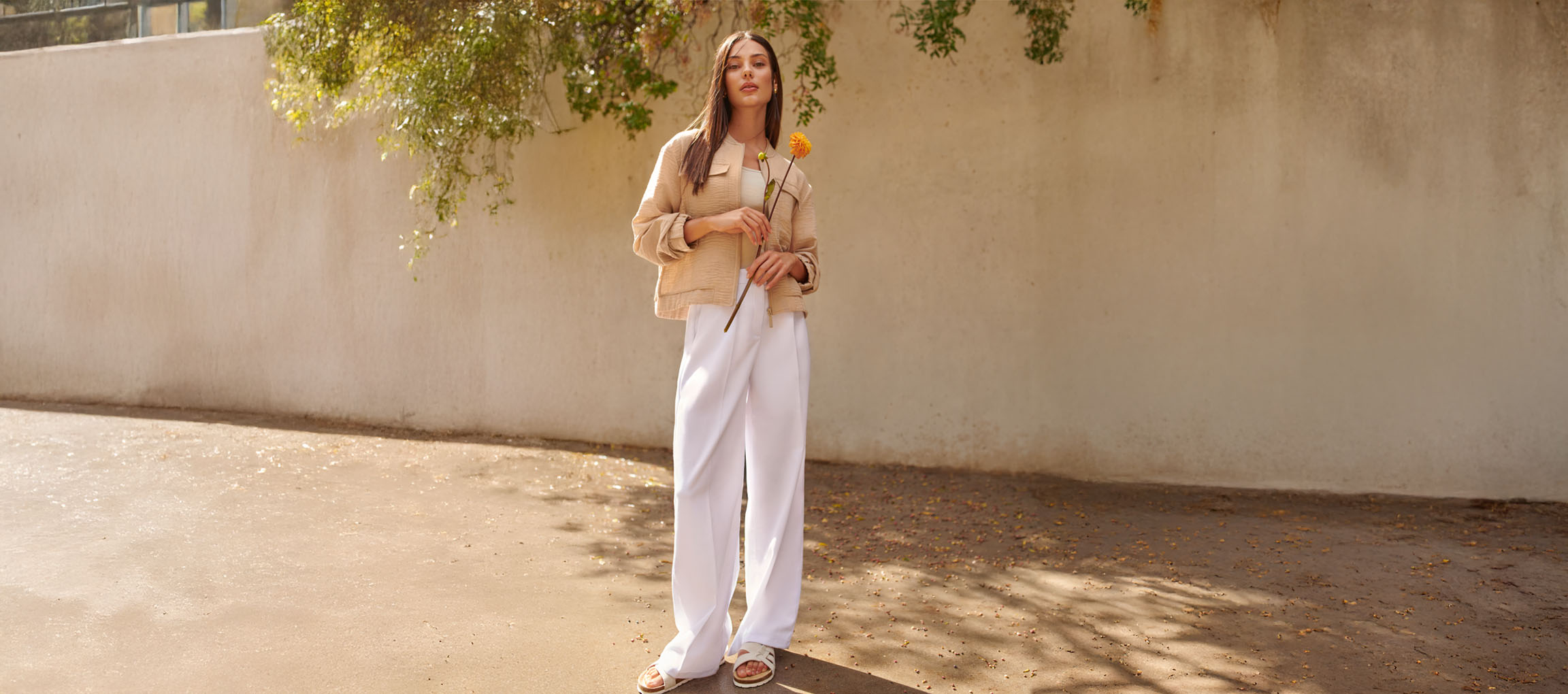 Kobieta w beżowej kurtce, białych spodniach i sandałach, trzymająca żółty kwiat.