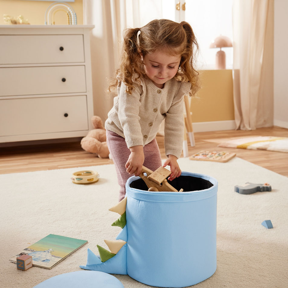 Dziecko wkłada drewniane zabawki do niebieskiego pojemnika na zabawki w kształcie dinozaura w sypialni.