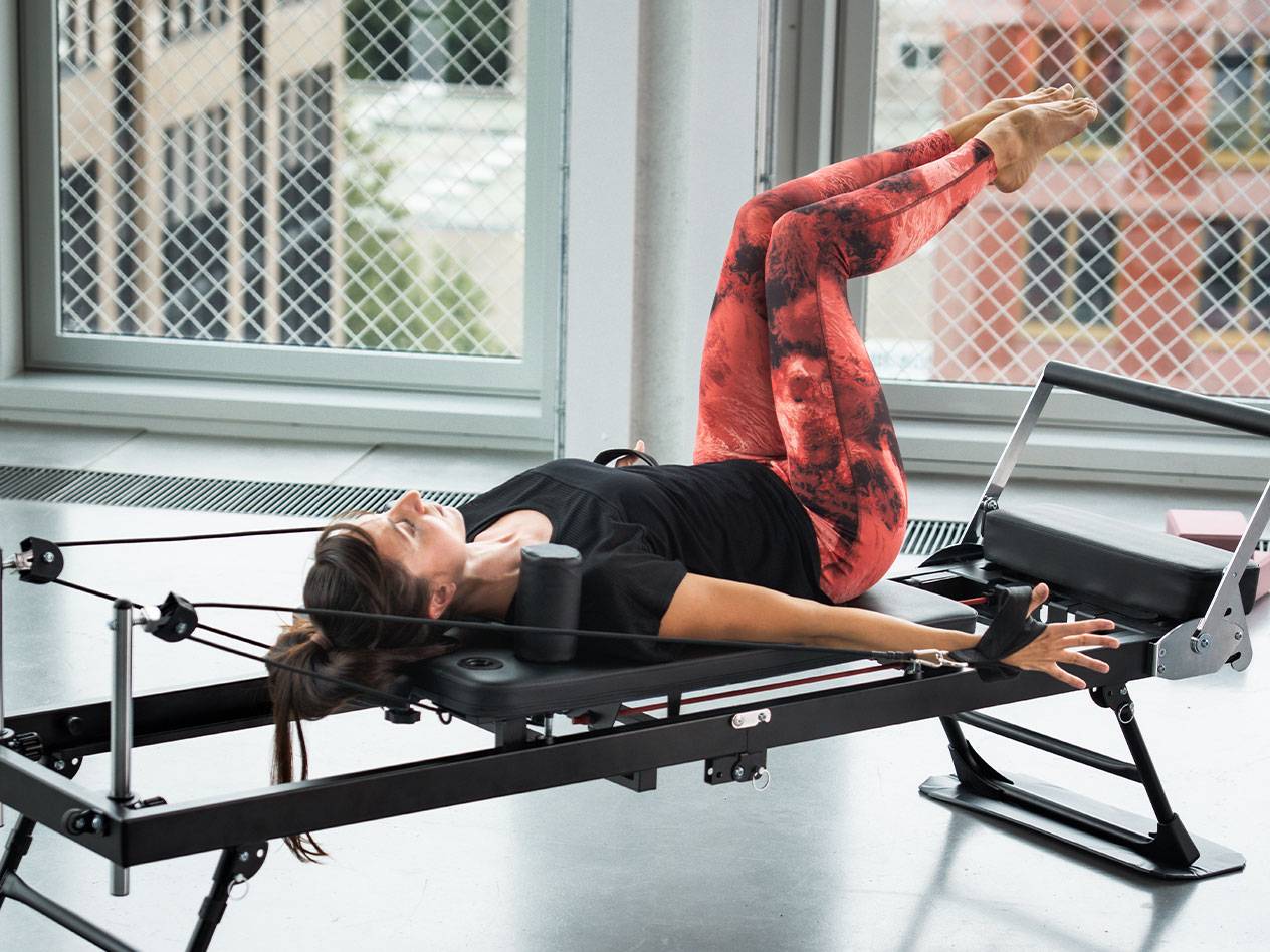 Kobieta ćwiczy Pilates na reformerze, w czerwono-czarnych legginsach